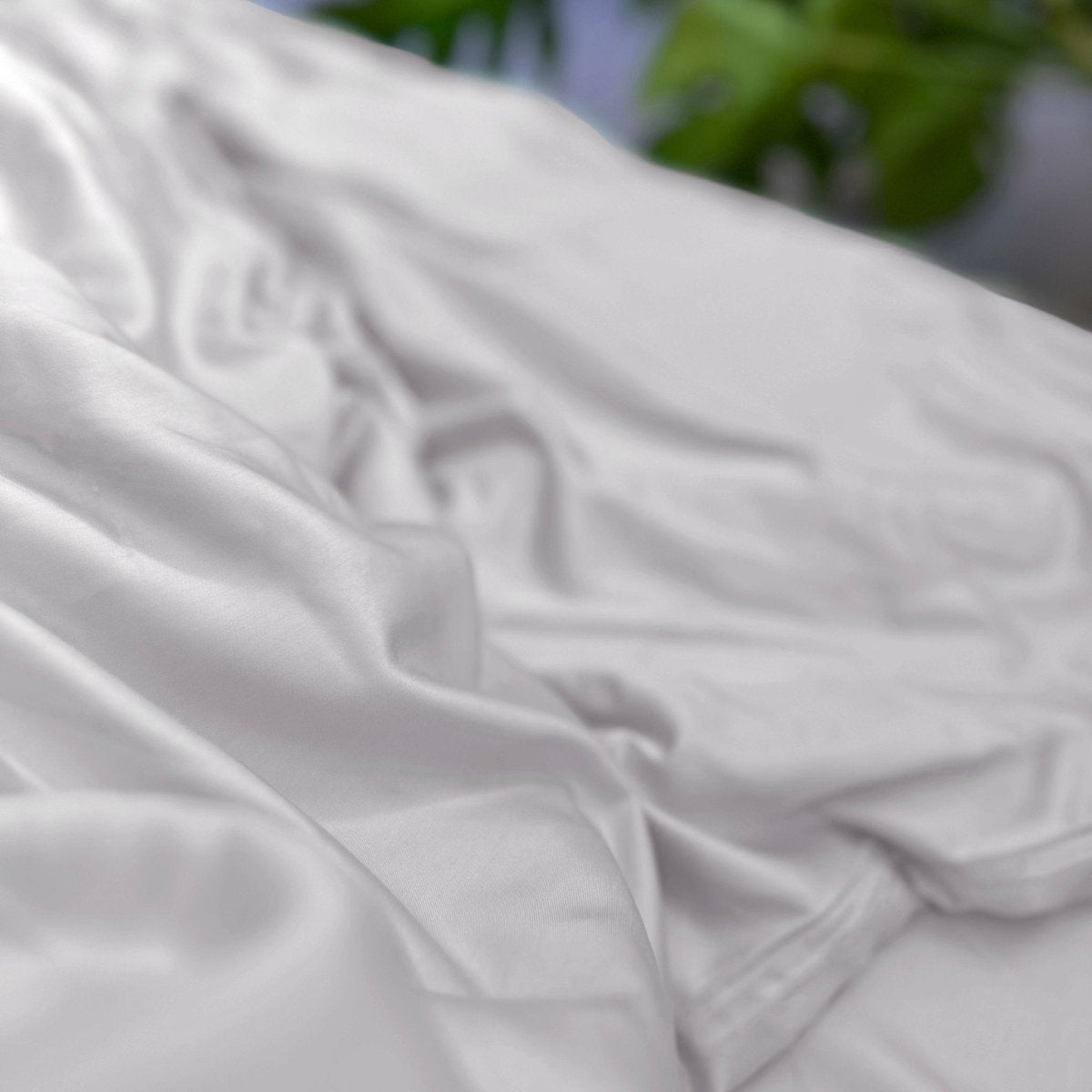 Lençol de algodão branco dobrado mostrando textura macia e detalhe de qualidade de tecido com fundo de folhagem verde