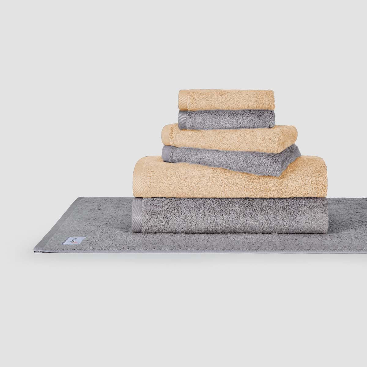 Stacked beige and gray towel set by Momo Cotton displayed on gray bath mat against white background
