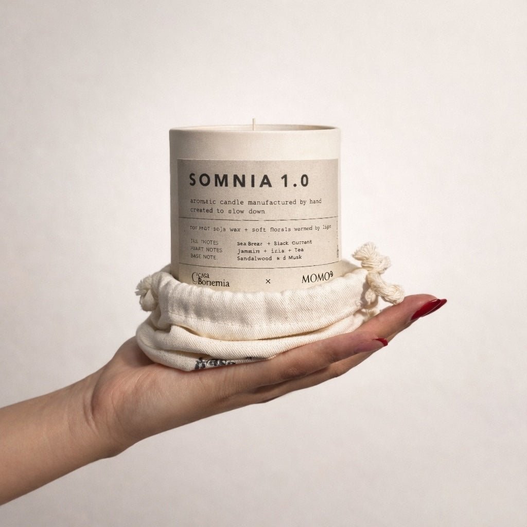 Mão segurando vela aromática Somnia 1.0 cor creme envolvida em tecido de algodão natural sobre fundo branco
