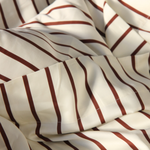 Lençol de cima em bambu acetinado creme com riscas bordeaux, dobrado e texturizado, mostrando o padrão listado e o acabamento