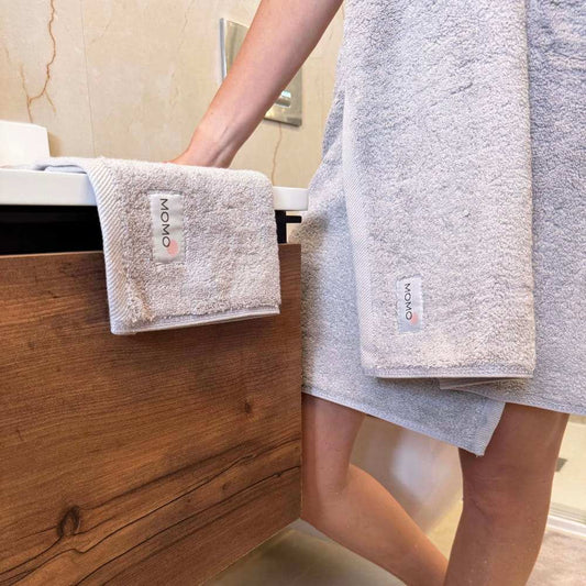 Conjunto de toalhas Momo em algodão exibido em prateleira de banheiro de madeira com pessoa segurando toalhas bege dobradas