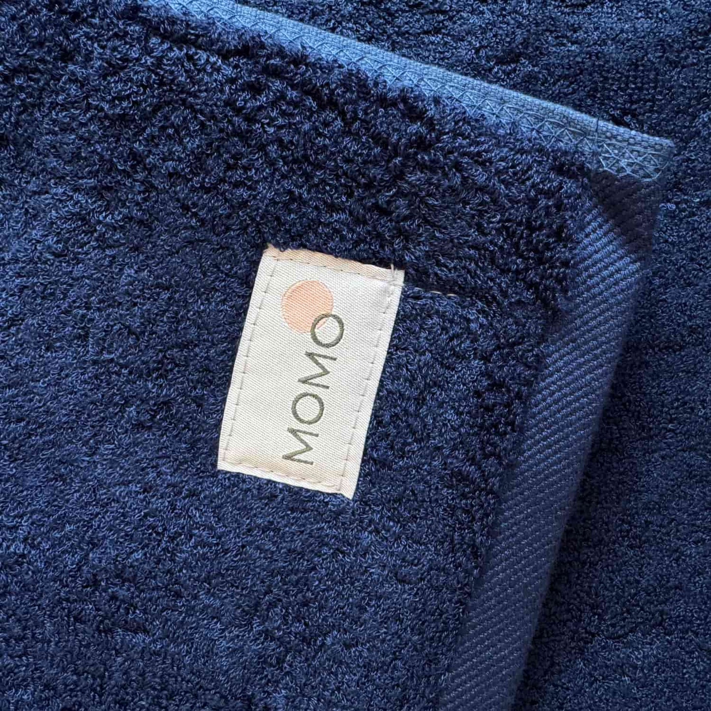 Tapete de banho azul marinho Wabi-sabi by Momo Cotton com etiqueta de marca visível na superfície texturizada