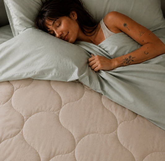 Mulher a dormir pacificamente em cama com colcha acolchoada bege e lençóis cinza claro da Momo Cotton