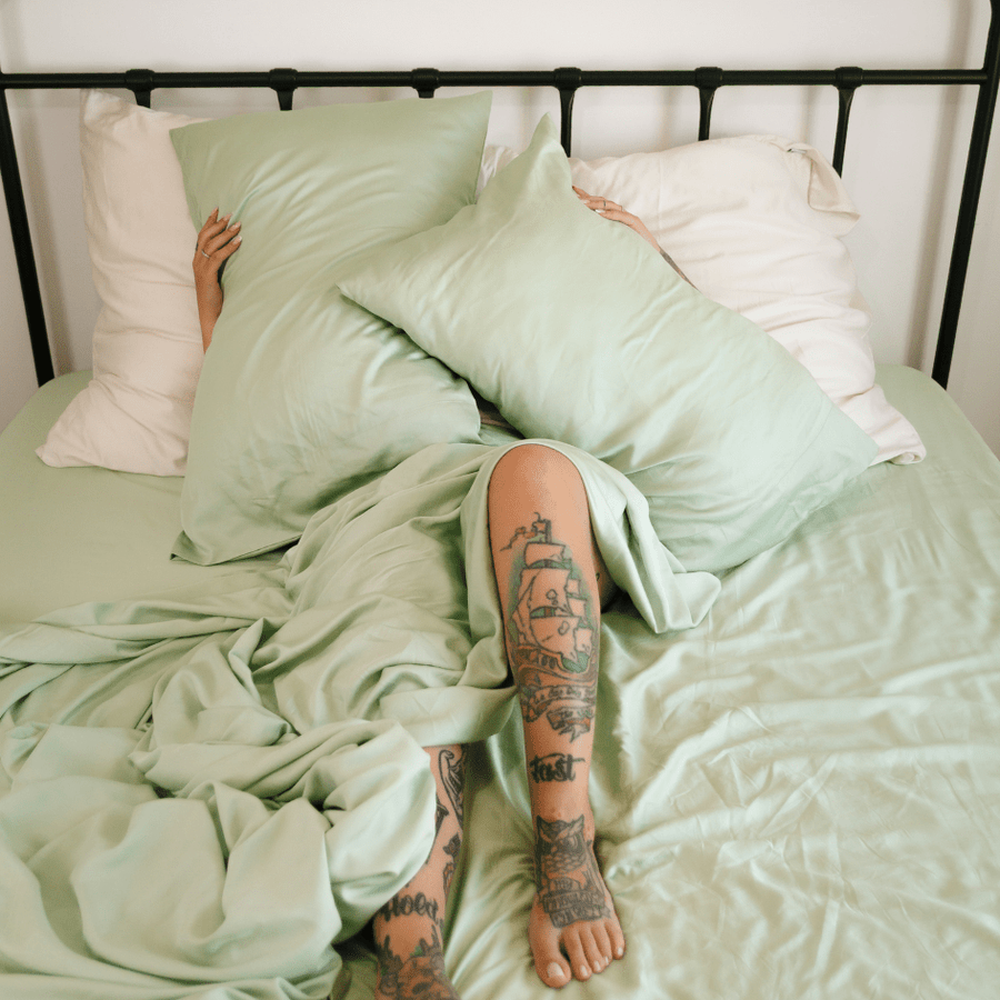 Pessoa deitada na cama usando conjunto de roupa de cama de bambu verde e branco com tatuagens visíveis nas pernas