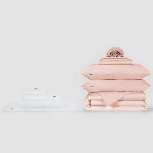 Conjunto de toalhas brancas e jogo de cama rosa com almofada decorativa apresentados em fundo neutro pela Momo Cotton