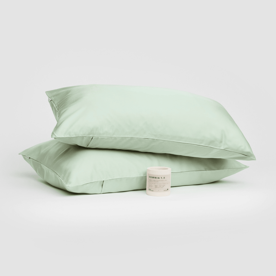 Par de fronhas de algodão verde sálvia empilhadas com vela aromática Somnia 1.0 ao fundo branco