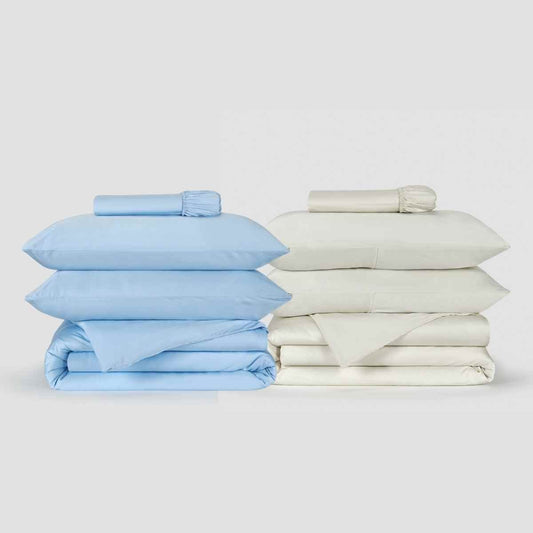 Dois conjuntos de capa de edredão Momo Cotton exibidos lado a lado, um azul claro e outro creme, com lençóis dobrados e rolos de linhas